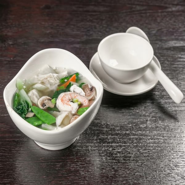Wonton soup with shrimp, mushrooms, and vegetables in a white bowl on a dark wooden table.