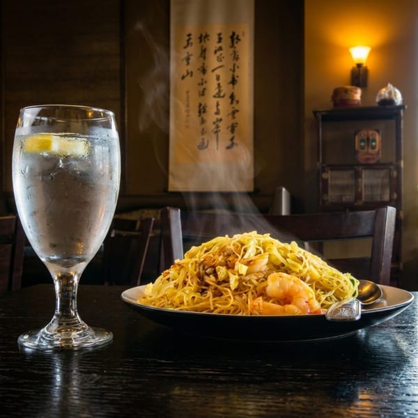 Steaming hot shrimp lo mein and a glass of water with lemon on a dark wooden table.